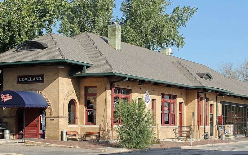The 1902 Colorado and Southern Railway depot on Railroad Avenue, a long tan-brick building with a hipped roof, green trim and a LOVELAND nameboard, now used by local businesses, on a sunny September morning.