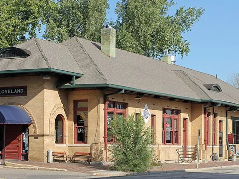 The 1902 Colorado and Southern Railway depot on Railroad Avenue, a long tan-brick building with a hipped roof, green trim and a LOVELAND nameboard, now used by local businesses, on a sunny September morning.