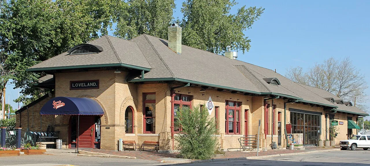 The 1902 Colorado and Southern Railway depot on Railroad Avenue, a long tan-brick building with a hipped roof, green trim and a LOVELAND nameboard, now used by local businesses, on a sunny September morning.