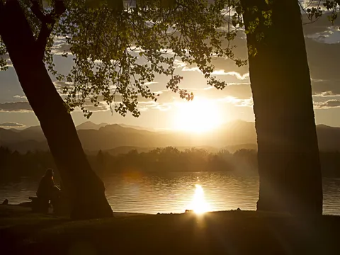 Sunset over Lake Loveland, framed by two trees on the shore, with a person sitting in silhouette at the water and the mountains beyond